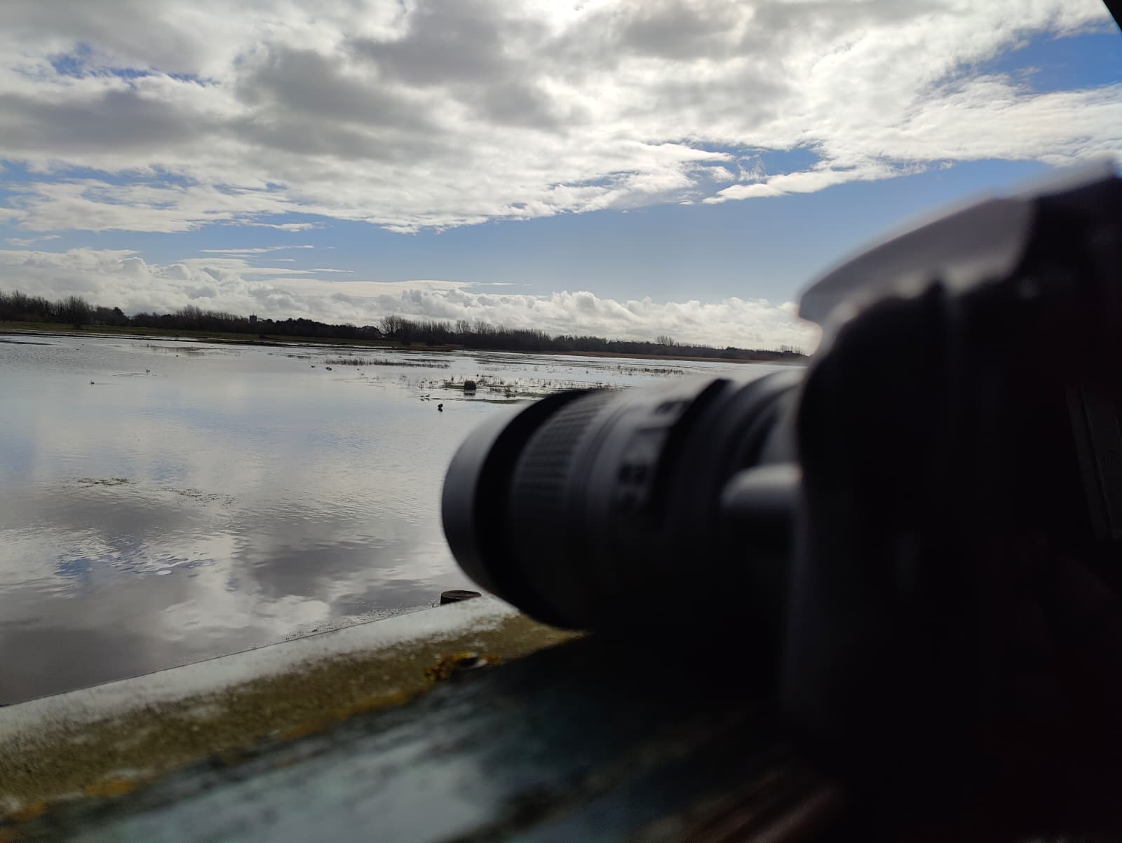 Camera on the window ledge of Nel's Hide at Marshside RSPB, pointed at the flooded scrape — clouds reflected in still water